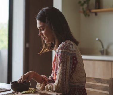 Woman in patterned dress enjoying breakfast at home near a sunlit window.