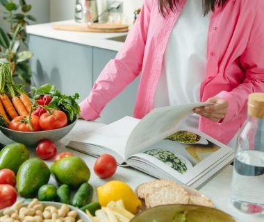 A person browsing a cookbook surrounded by fresh vegetables in a bright kitchen setting.